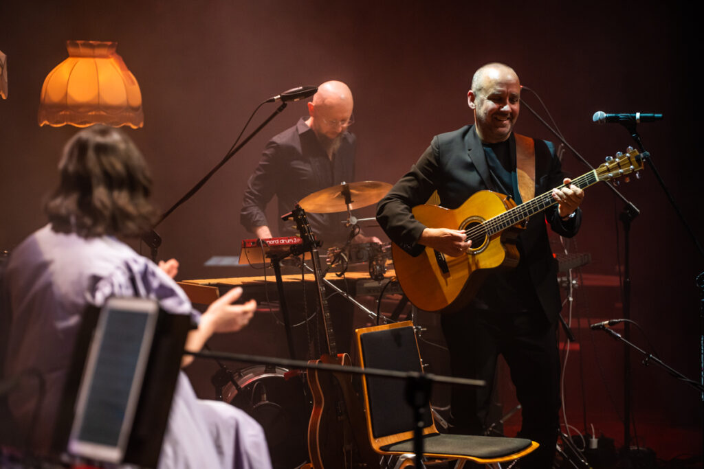 Koncert. Mężczyzna w garniturze gra na gitarze akustycznej. W tle mężczyzna gra na klawiszach. Przed nimi siedzi kobieta, która klaszcze.