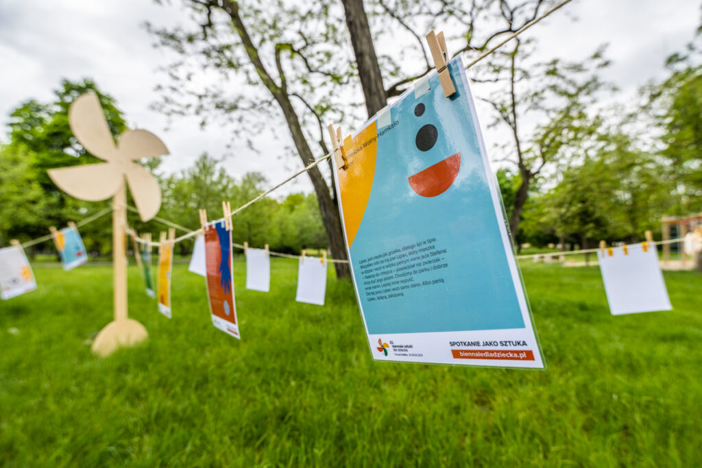 W parku instalacja złożona z dużego drewnianego wiatraka, do którego są przywiązane trzy sznurki. Na końcu każdego sznurka znajduje się wbity w ziemię palik. Na sznurkach jak pranie rozwieszone są teksty wierszy.