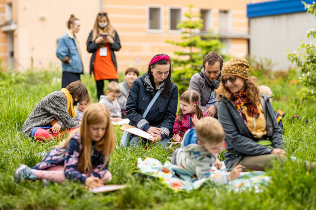 Słoneczny dzień. Na bujnej trawie, w kilku grupach, siedzą dzieci i dorośli. Pochyleni są nad kartkami.