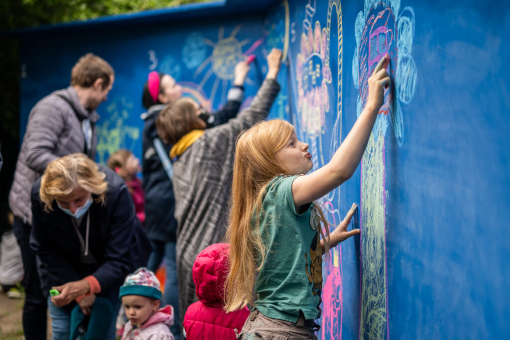 Dzieci i dorośli malują na ścianach kolorowe rysunki. Na pierwszym planie dziewczynka maluje drzewo.
