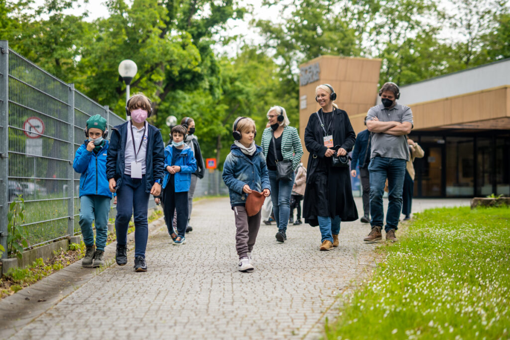 Grupa dzieci i dorosłych osób przed budynkiem Sceny Wspólnej. Uśmiechają się, na uszach mają słuchawki, idą na spacer.