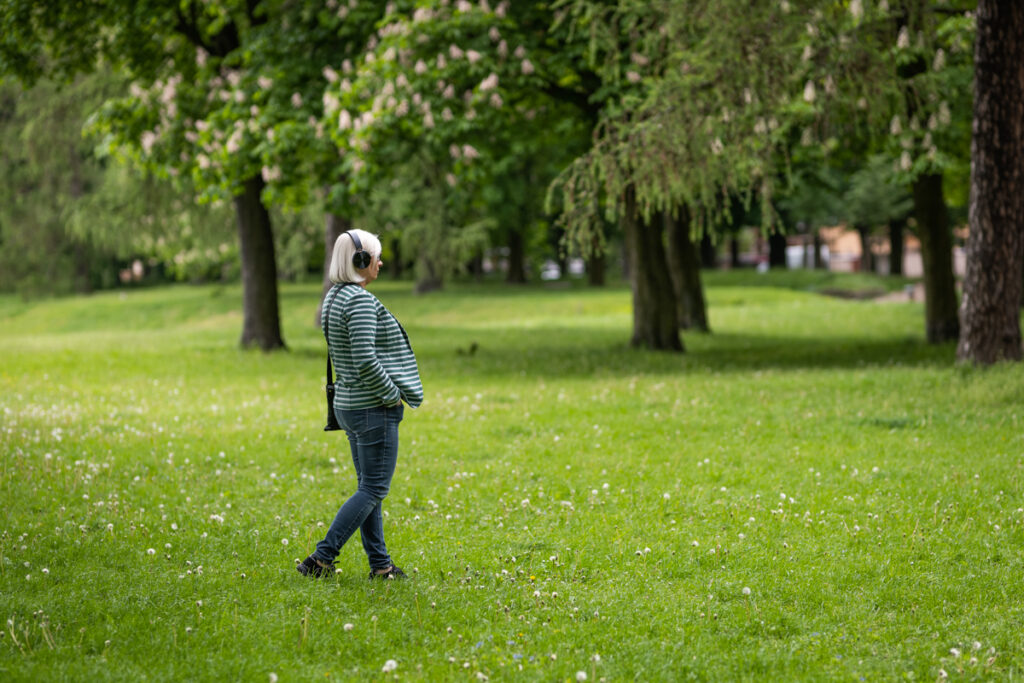 W parku na polanie, bokiem do osoby robiącej zdjęcie, stoi kobieta z słuchawkami na uszach. Patrzy z zainteresowaniem przed siebie.