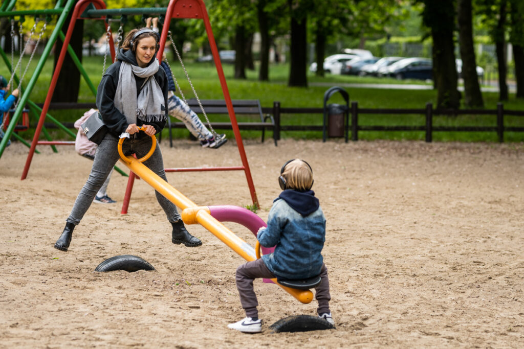 Plac zabaw w parku. Kobieta i dziecko bawią się na huśtawce. Mają na uszach słuchawki.