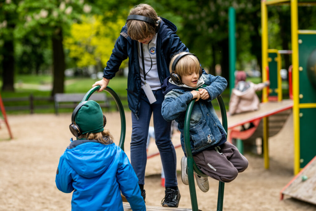 Czworo dzieci na placu zabaw w parku. Każde z nich ma słuchawki na uszach.