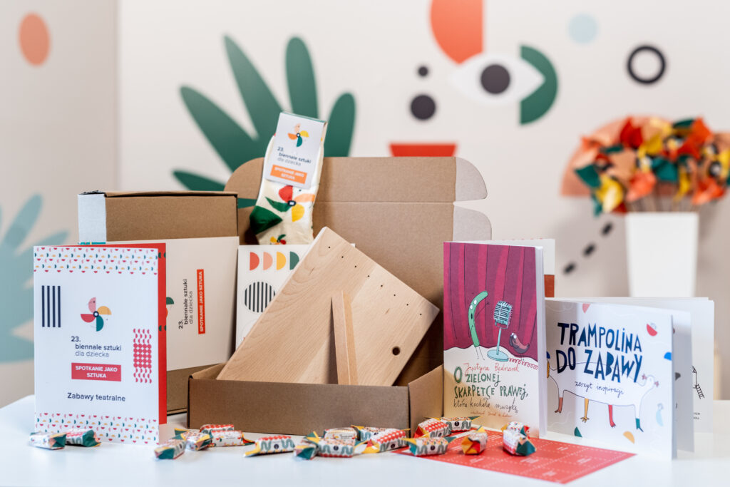 Na stole kolorowe elementy wchodzące w skład pakietu Biennale na wynos - zeszyty zabaw, publikacja z opowiadaniem, drewniana cytra, cukierki 