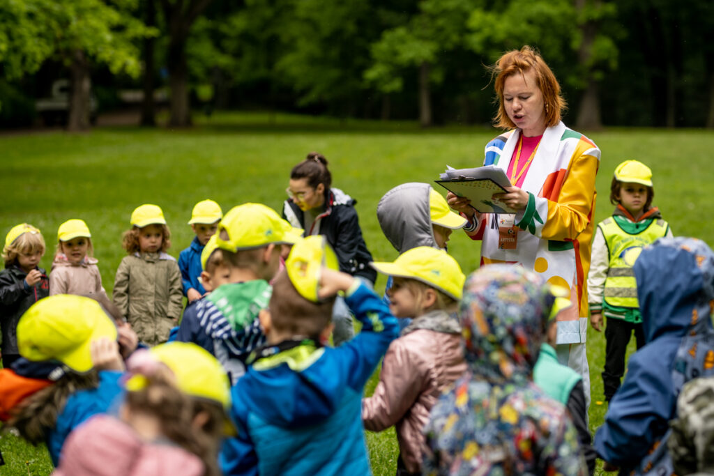 Park. Młoda kobieta w kolorowym kostiumie-garniturze otoczona przez grupę dzieci w wieku przedszkolnym. Trzyma w dłoniach kartki, czyta. Za nimi drzewa.