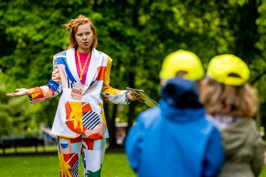 Park. Młoda kobieta w kolorowym kostiumie-garniturze mówi do dwójki dzieci.