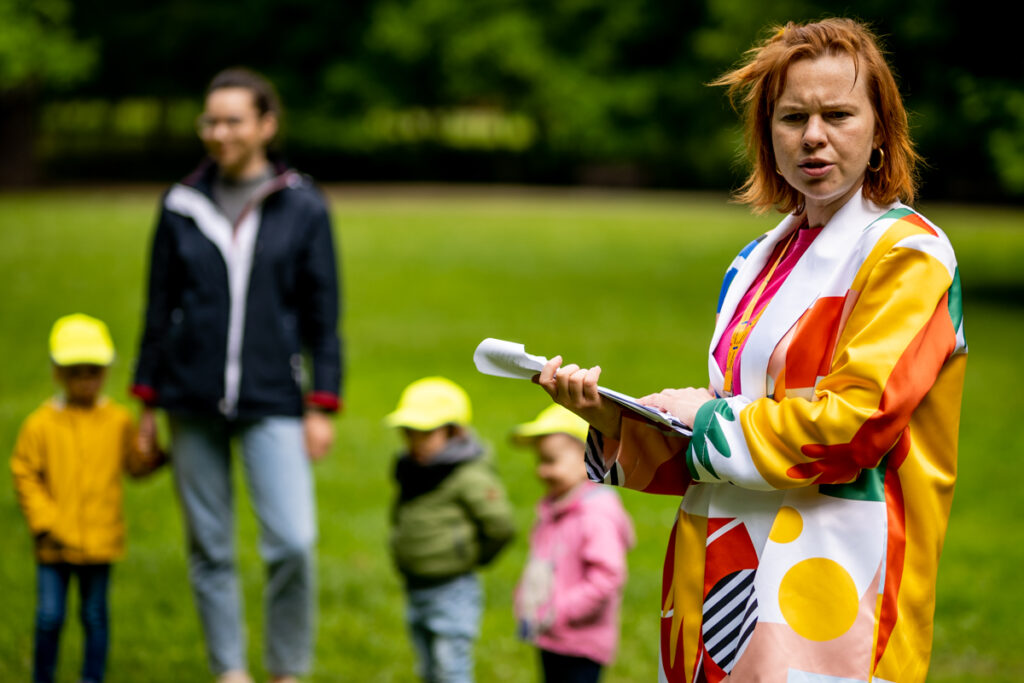 W parku kobieta ubrana w kolorowy kostium przypominający garnitur trzyma w dłoniach stos kartek, czyta. Tuż za nią dzieci w wieku przedszkolnym oraz mężczyzna - słuchają, tworząc publiczność.