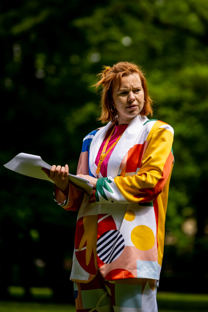 Młoda kobieta w kolorowym kostiumie trzyma w dłoniach kartki, czyta. Świeci słońce, na drugim planie drzewa.