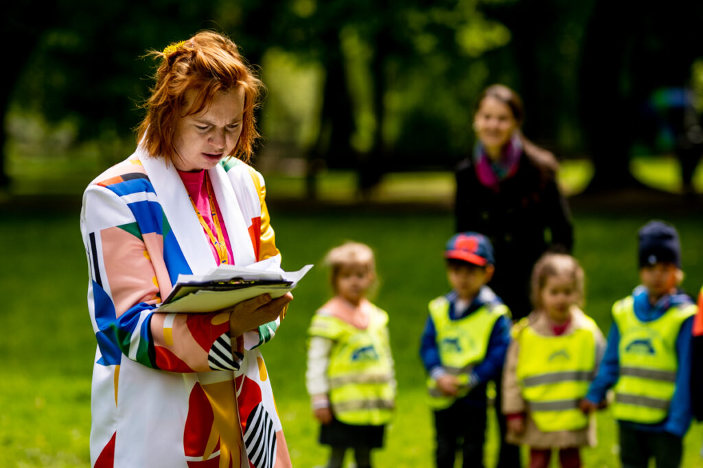 W parku kobieta ubrana w kolorowy kostium przypominający garnitur trzyma w dłoniach stos kartek, czyta. Tuż za nią dzieci w wieku przedszkolnym oraz kobieta - słuchają, tworząc publiczność.