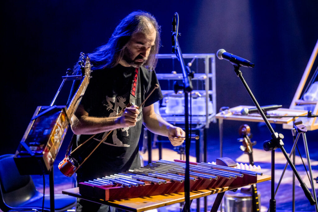 Koncert. Mężczyzna z długimi włosami uderza pałkami w dzwonki. Przed nim stoją mikrofony.