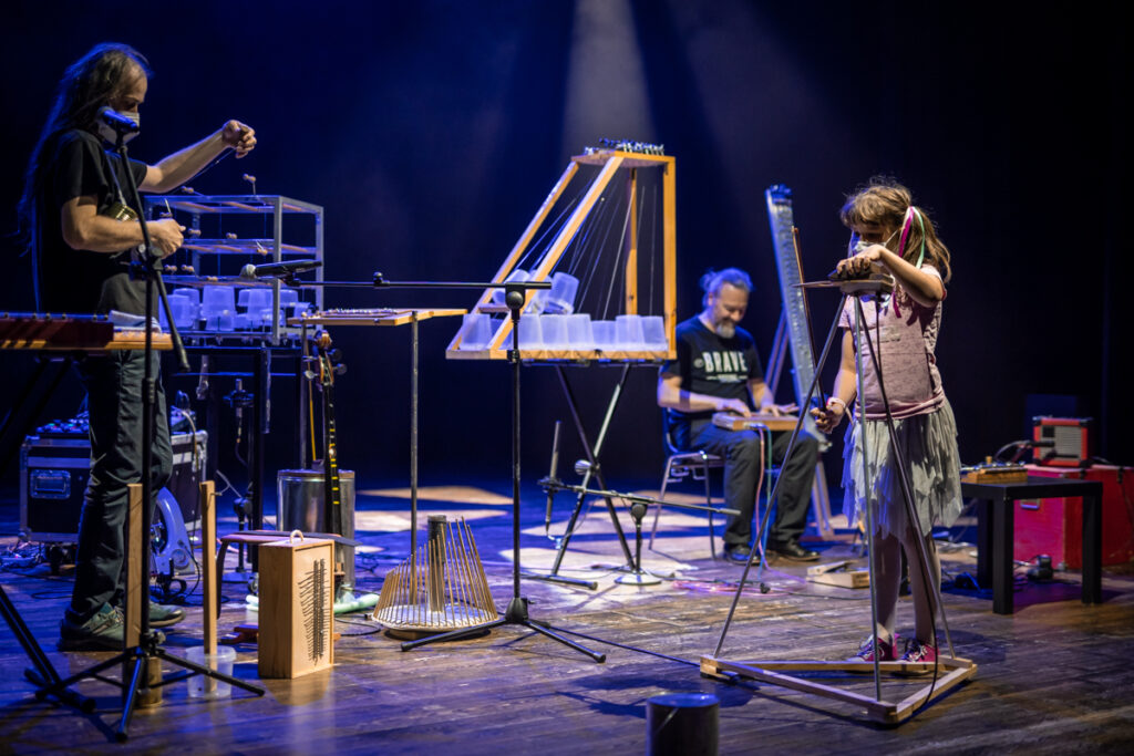 Koncert. Na scenie znajduje się wiele niekonwencjonalnych instrumentów. Dziewczynka gra na konstrukcji przypominającej ostrosłup.