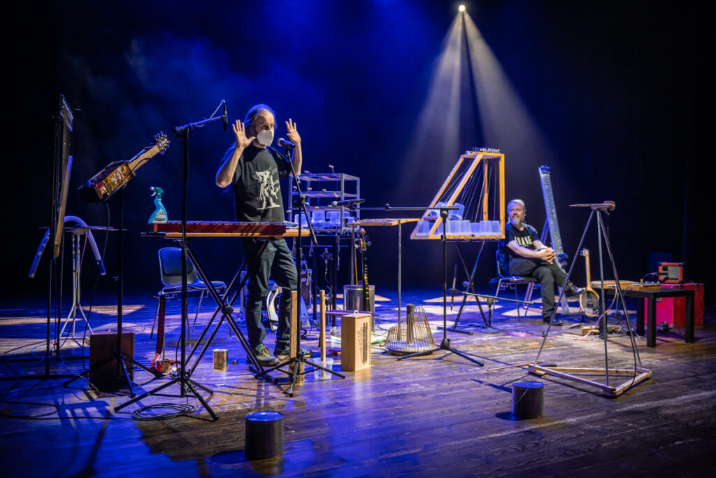 Koncert. Scena wypełniona niekonwencjonalnymi instrumentami. Mężczyzna w maseczce śpiewa do mikrofonu. Gestykuluje. W tle inny mężczyzna siedzi na krześle i słucha.