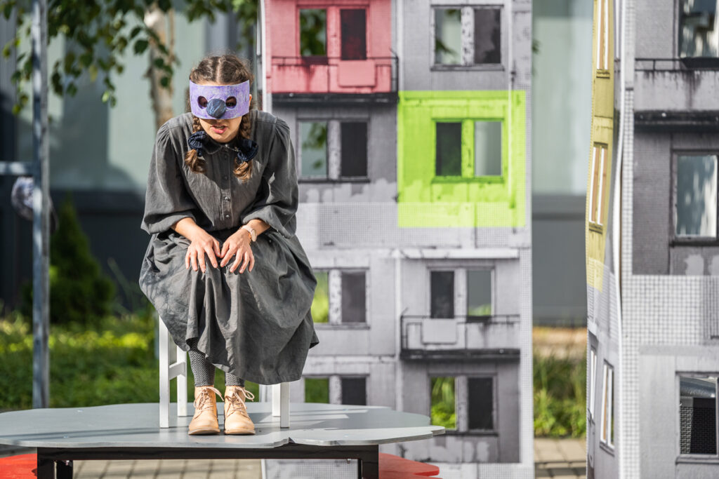 Na krześle siedzi kobieta w masce ptaka. Ubrana jest w szary kostium. Pochyla się w kierunku widowni. W tle scenografia przypominająca bloki mieszkalne.