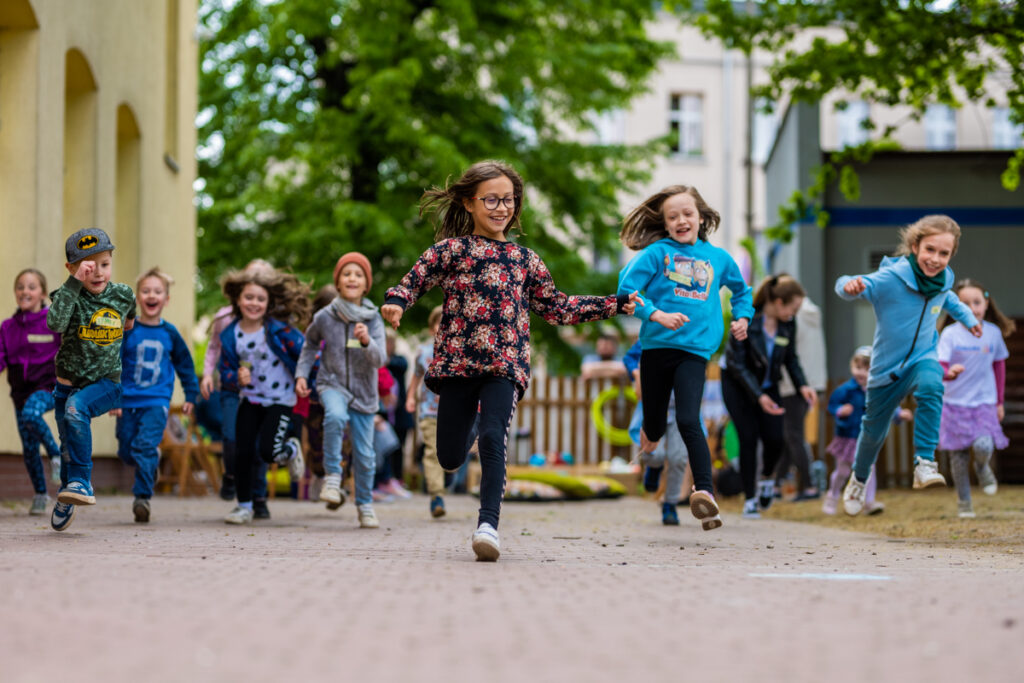 Podwórko. Grupa uśmiechniętych dzieci biegnie. Wyścig prowadzi dziewczynka w okularach.