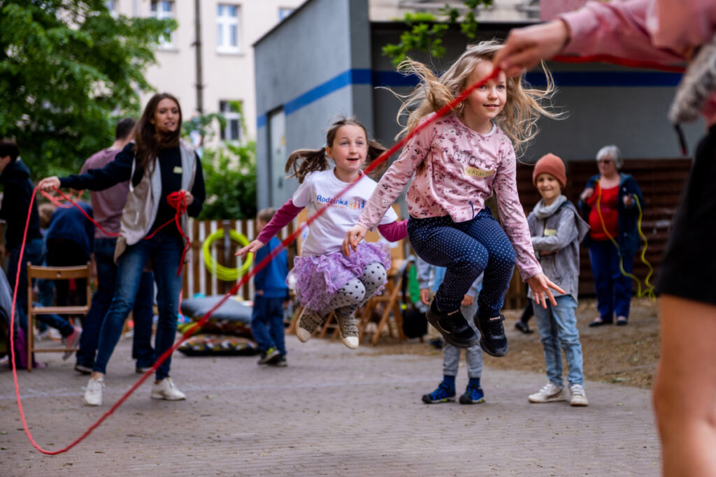 Podwórko. Dziewczynki skaczą n a skakance, którą kręcą dwie osoby.