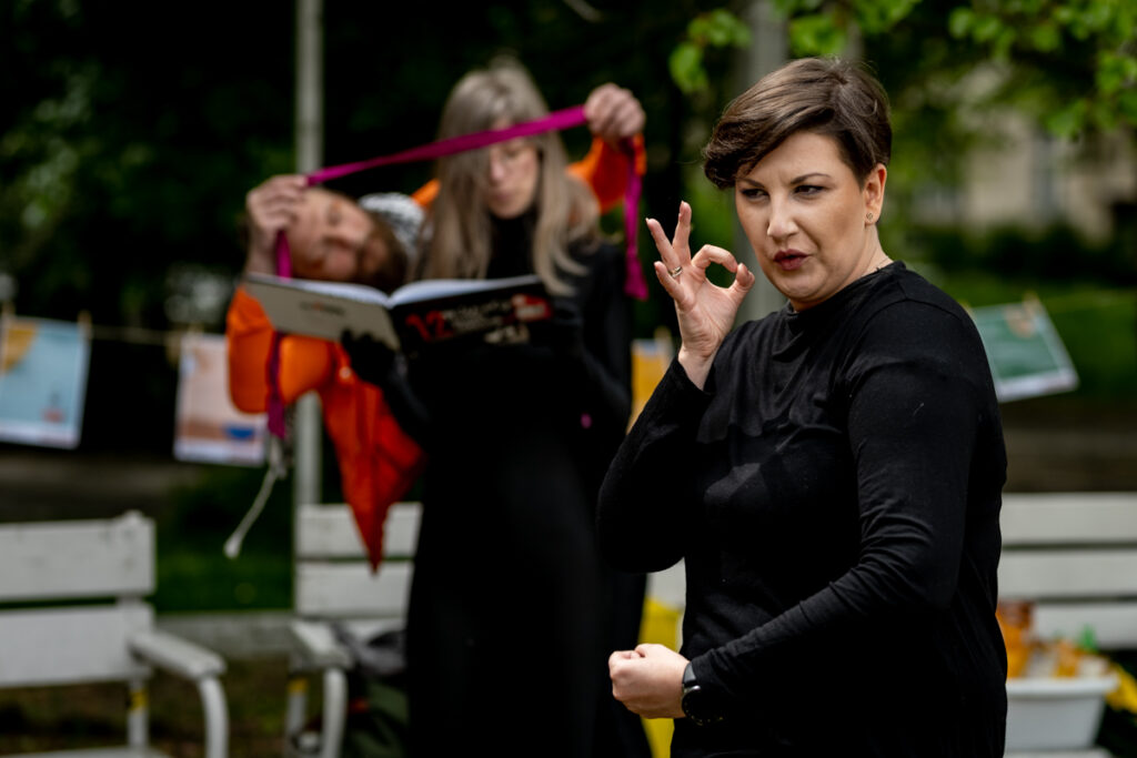 Park. Na pierwszym planie kobieta w czerni gestykuluje. W tle kobieta trzymająca książkę, czyta. Za nią mężczyzna w pomarańczowej kurtce. Trzyma w dłoniach czerwoną wstążkę, którą próbuje zawiązać na głowie kobiety. Dookoła białe ławki.