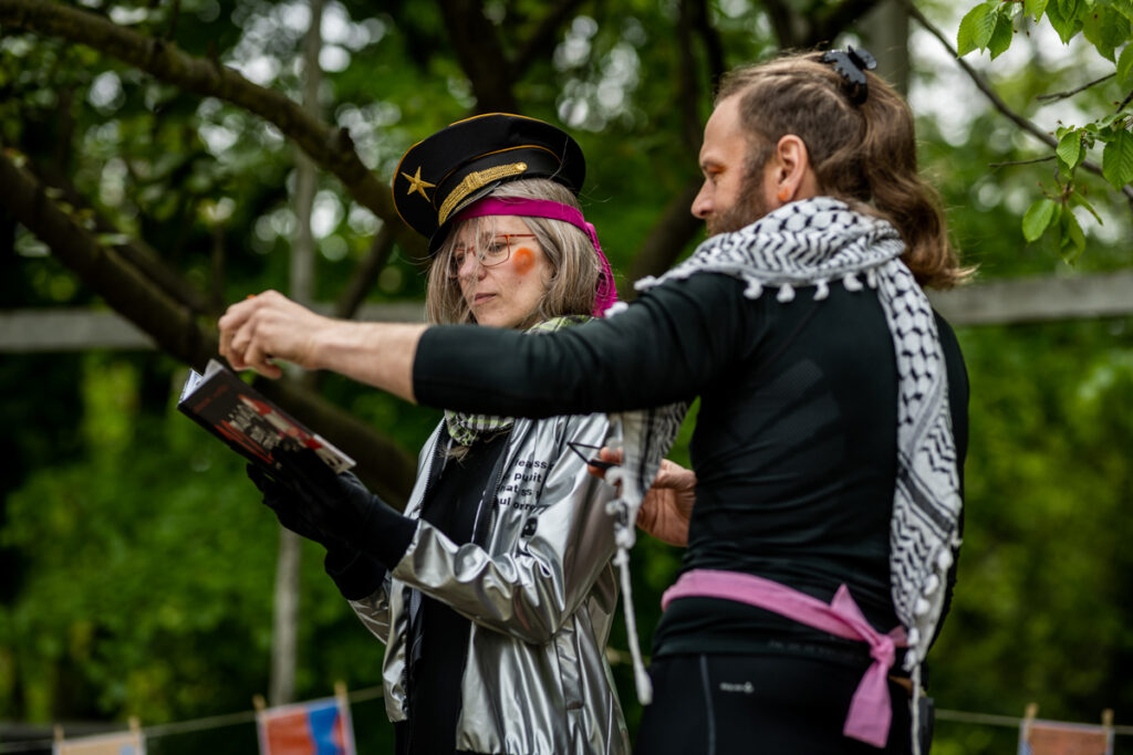 Park. Na pierwszym planie kobieta czytająca książkę. Obok niej stoi mężczyzna, który wyciąga rękę, by przewrócić kartkę.