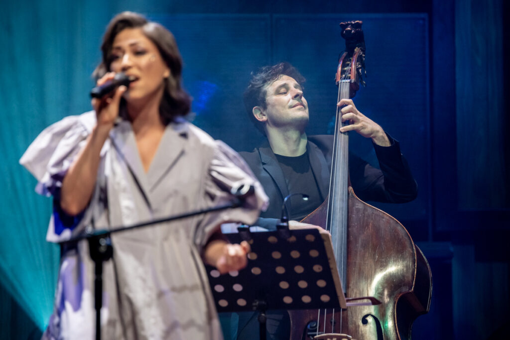 Koncert. Wokalistka śpiewa do mikrofonu. W tle mężczyzna gra na kontrabasie.