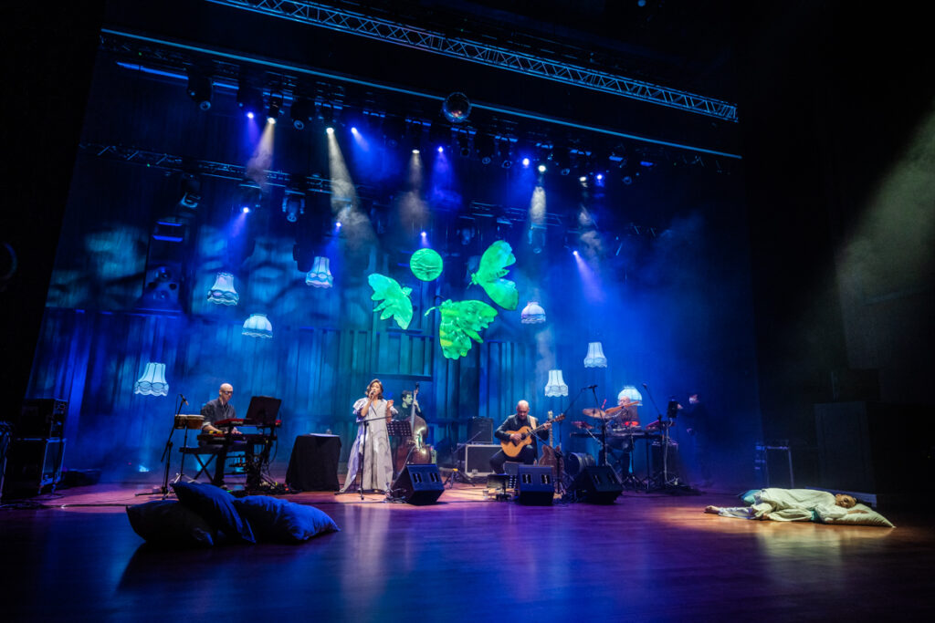 Koncert. W głębi sceny wokalistka i muzycy. Nad nimi zwisają abażury oraz scenografia przypominająca ogromne ćmy. Przed zespołem dwie osoby leżą na poduszkach.