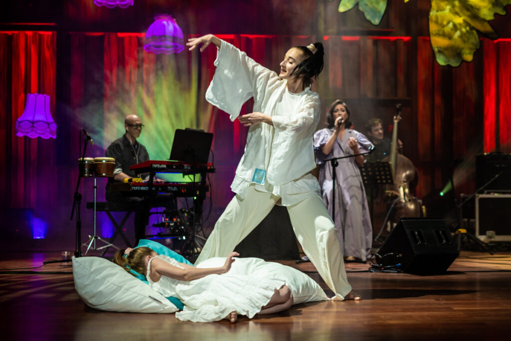 Koncert. Dziewczyna w sukni leży na poduszkach. Nad nią stoi tancerka i porusza rękami. W tle śpiewająca wokalistka i zespół muzyków. Nad sceną zwisają abażury.