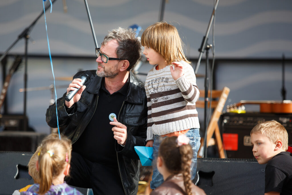 Na zdjęciu mężczyzna, a obok niego chłopiec. Oboje stoją na scenie i patrzą w lewo. Mężczyzna trzyma w dłoni mikrofon, a chłopiec niebieską papierową łódkę.