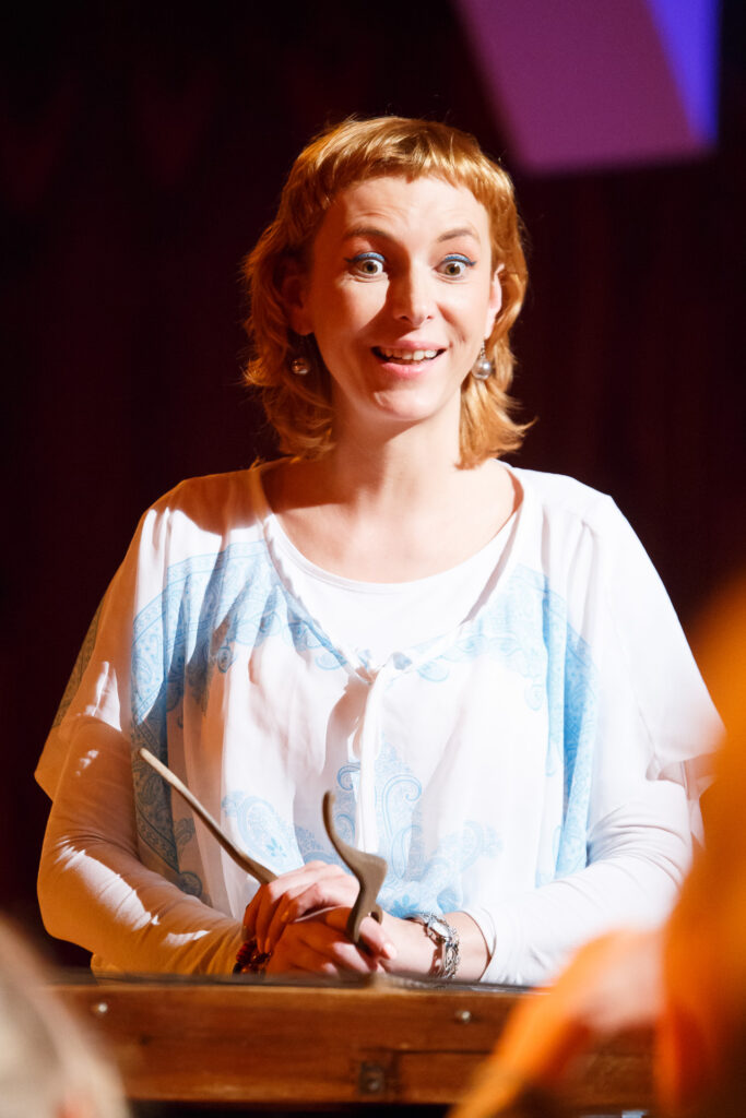 Kobieta stoi przed mównicą. Ubrana jest na biało i niebiesko. Robi zdziwioną minę i uśmiecha się szeroko.