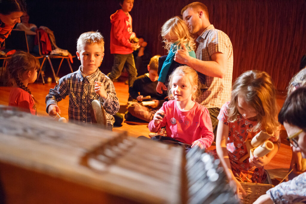Na zdjęciu dzieci bawiące się różnymi instrumentami. W tle tata z córką.