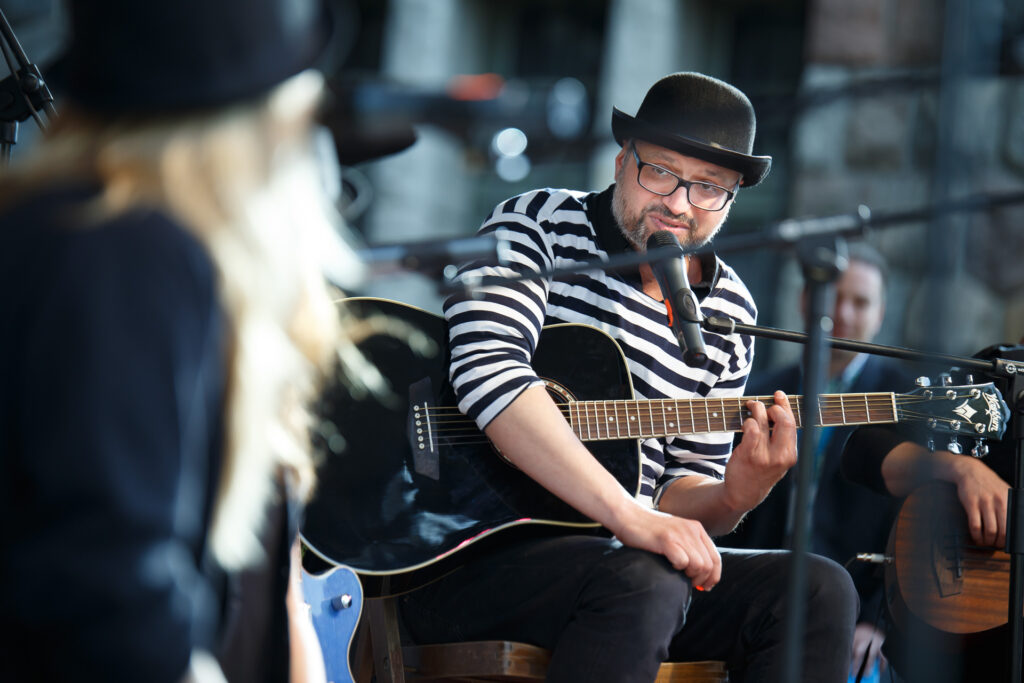 Na zdjęciu mężczyzna siedzi na krześle na scenie. Gra na gitarze i śpiewa do mikrofonu. Ubrany jest w czarne spodnie, koszulkę w białe i czarne paski oraz czarny kapelusz. Mężczyzna ma brodę i okulary.