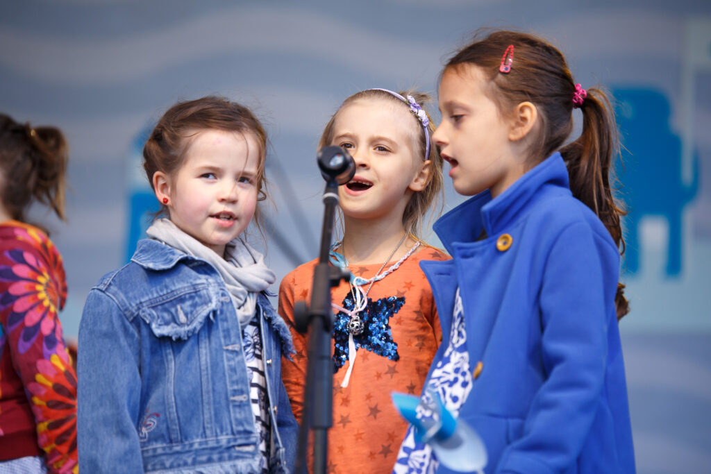 Na zdjęciu trzy dziewczynki ubrane na niebiesko i pomarańczowo. Wszystkie stoją na scenie i śpiewają razem do jednego mikrofonu.