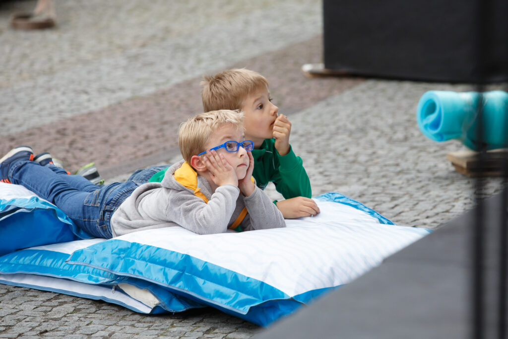 Na zewnątrz. Dwaj chłopcy leżą na brzuchach na poduszkach na betonie i patrzą przed siebie