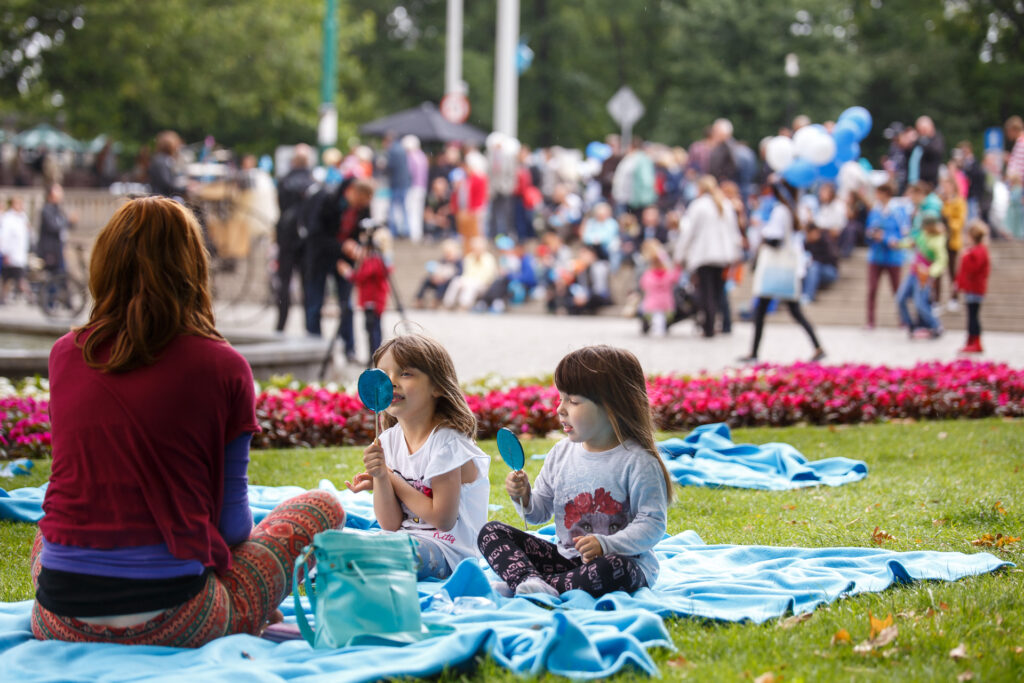 Na zewnątrz. Na niebieskich kocach na trawie siedzą dwie dziewczynki i kobieta. Dziewczynki jedzą niebieskie lizaki w kształcie koła. W tle inni ludzie.