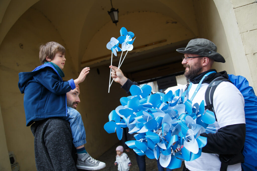 Na zewnątrz. Mężczyzna w kapeluszu z niebieskim plecakiem rozdaje niebiesko białe wiatraczki. Jednego wręcza chłopcu, który siedzi u taty na barana.