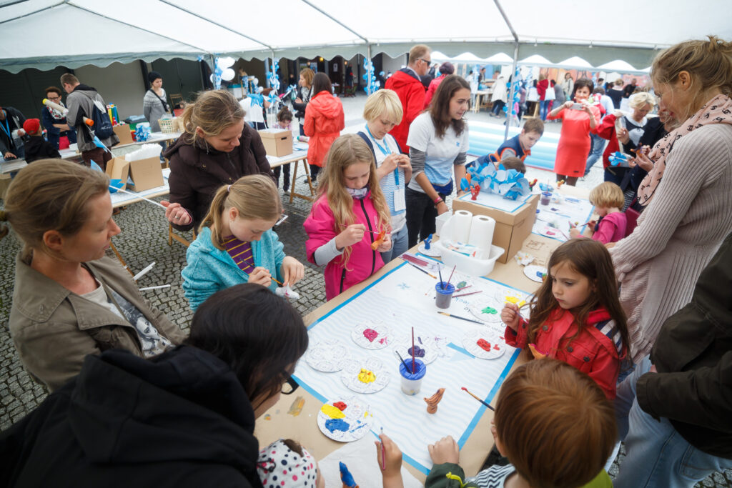 Dziedziniec CK Zamek. Na nim porozstawione namioty, a pod nimi ustawione stoły. Wokół stołów dzieci i dorośli. Na stołach farby, brystole i inne przybory plastyczne. Dzieci malują