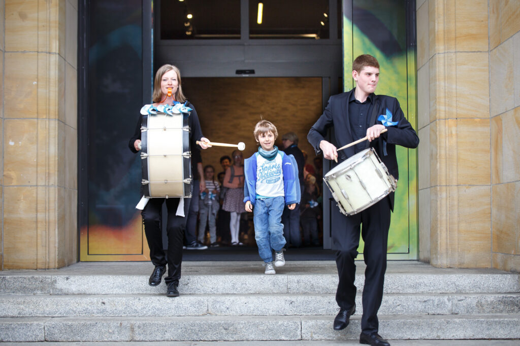 Na zdjęciu mężczyzna i kobieta. Grając na bębnach ozdobionych niebieskimi i białymi wstążkami wychodzą z CK Zamek. Między nimi chłopiec. Za nimi wychodzą też inne dzieci.
