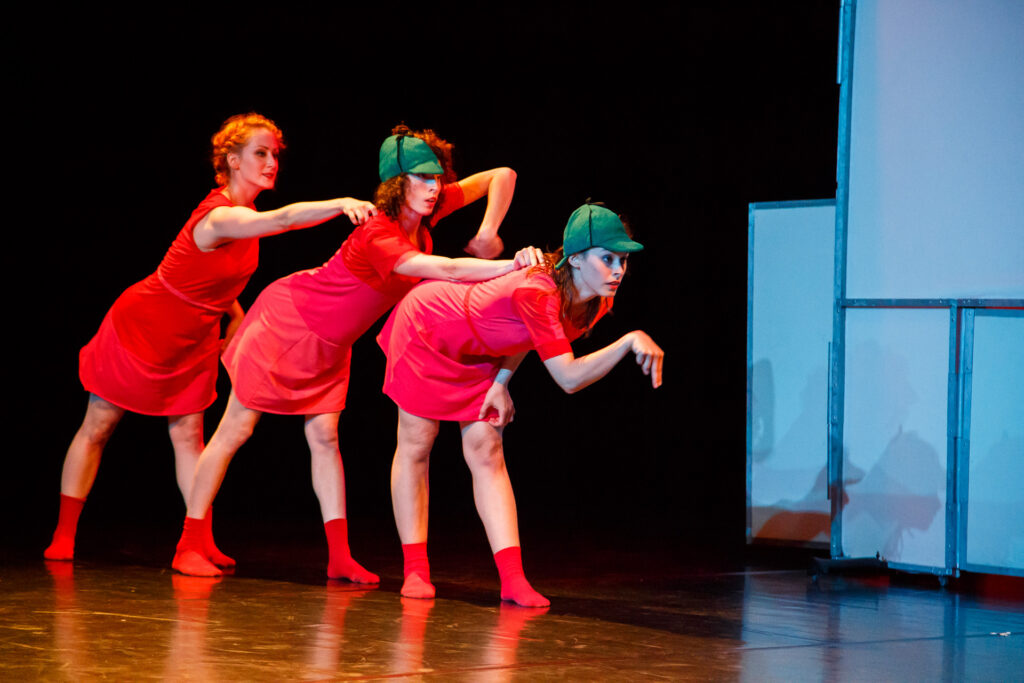 Na scenie trzy aktorki ubrane w czerwone sukienki i skarpetki. Stoją jedna za drugą nachylone i oparte na sobie. Dwie pierwsze mają ubrane zielone czapki z daszkiem.