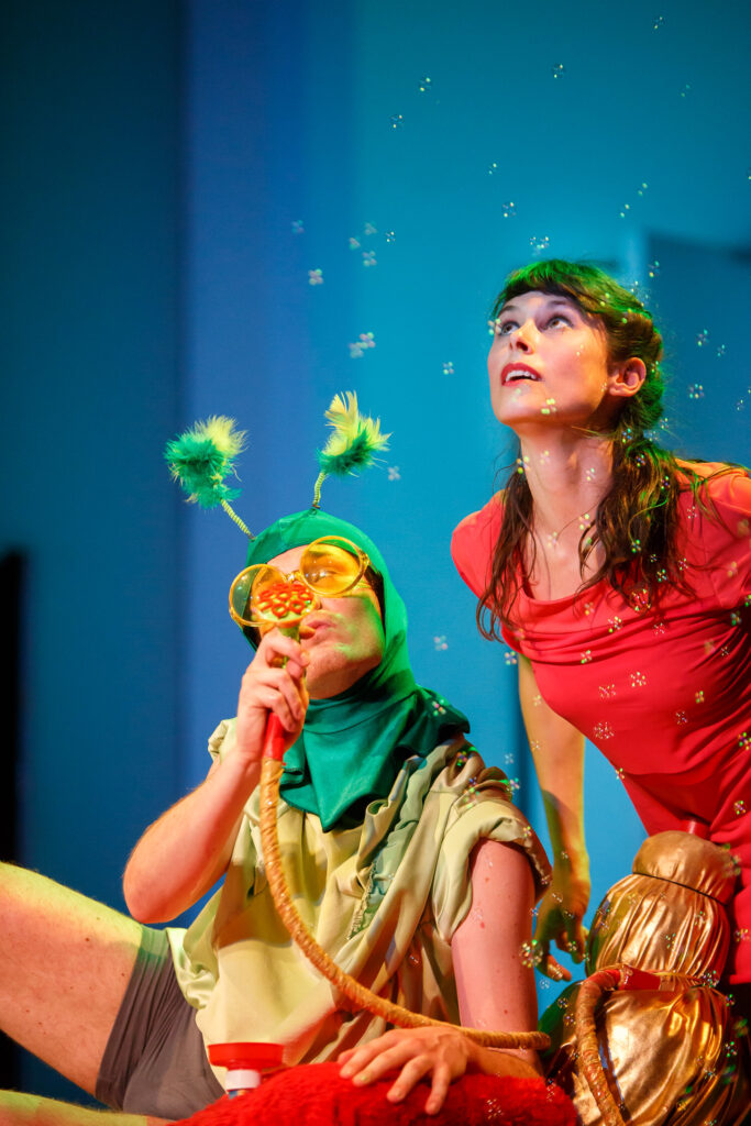 Na scenie dwoje aktorów. Mężczyzna ubrany w zieloną kominiarkę z czułkami i pomarańczowe okulary puszcza bańki. Kobieta w czerwonej sukience ma zdziwioną minę i obserwuje bańki.