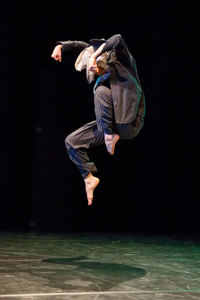 Aktor na scenie ubrany na czarno. Na zdjęciu uchwycony skulony w wysokim podskoku.