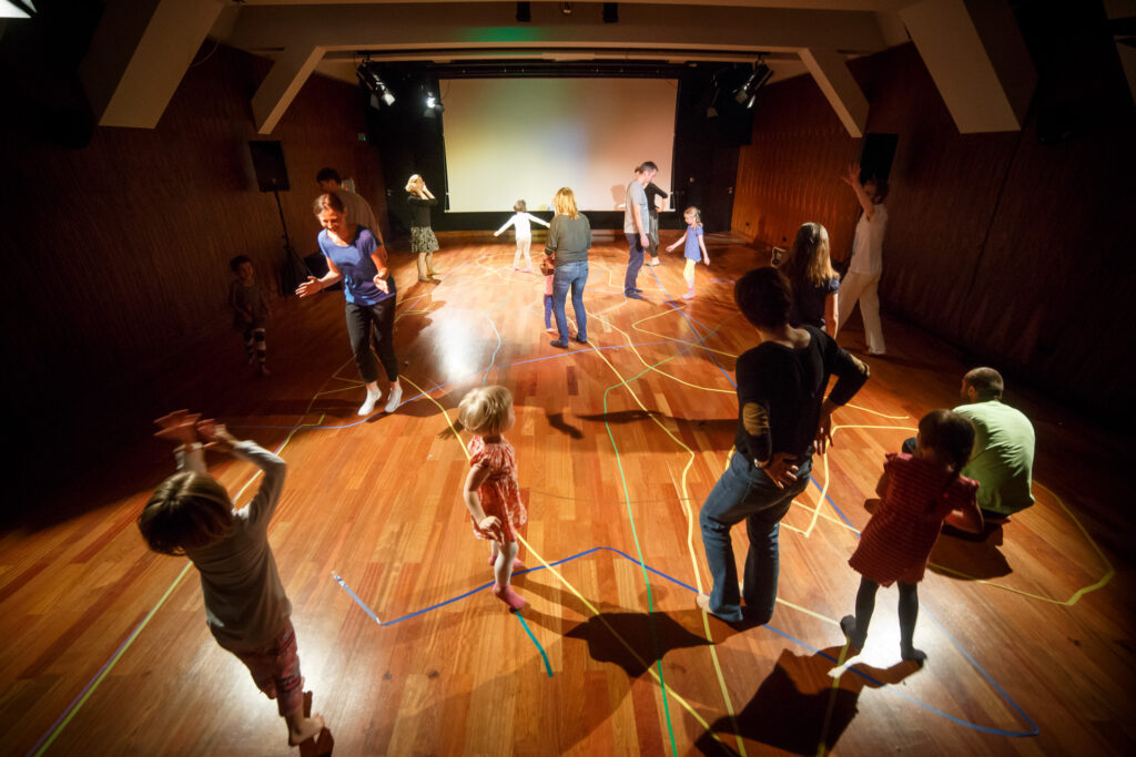 Grupa dzieci i dorosłych w sali. Chodzą po ścieżkach wyznaczonych na podłodze kolorowymi taśmami.