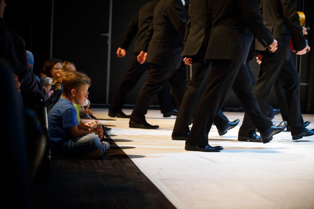 Na zdjęciu fragment widowni na granicy ze sceną. Dzieci obserwują grupę aktorów w czarnych garniturach, którzy maszerują w przeciwnym do nich kierunku.
