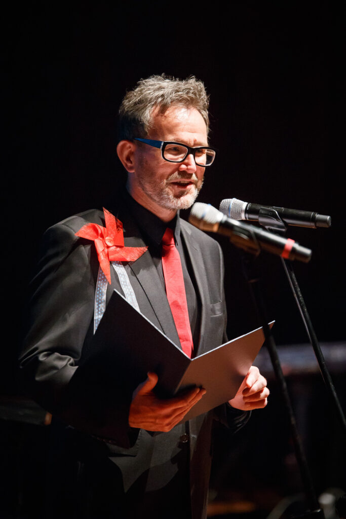 Na zdjęciu mężczyzna mówi do mikrofonu. Ubrany jest w czarny garnitur z czerwonym krawatem. Do piersi ma przytwierdzony czerwony wiatraczek z papieru. W dłoniach trzyma rozłożoną teczkę