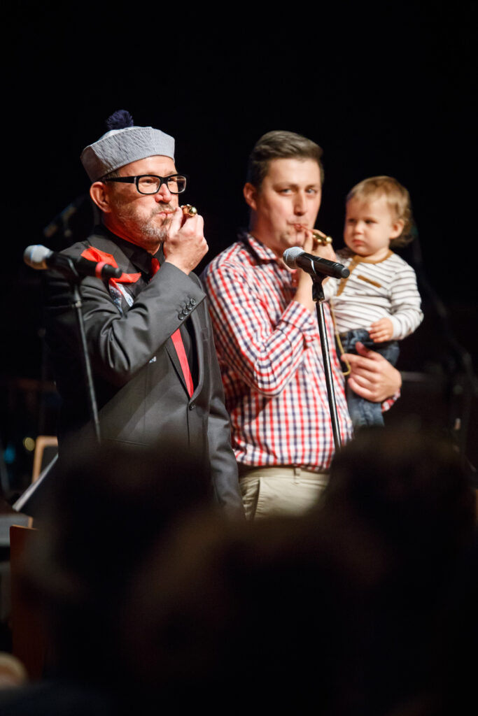 Na scenie występujący mężczyzna. Ubrany na czarno z czerwonym krawatem i czerwonym wiatraczkiem przytwierdzonym do piersi. Mężczyzna gra dmucha w instrument. Obok niego stoi mężczyzna z dzieckiem na rękach i naśladuje go.