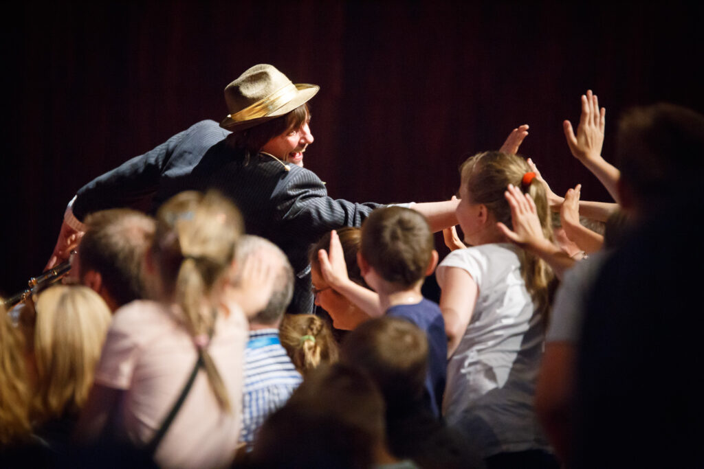 Na zdjęciu aktor w kapeluszu, ubrany na czarno przybija piątki z dziećmi z widowni.