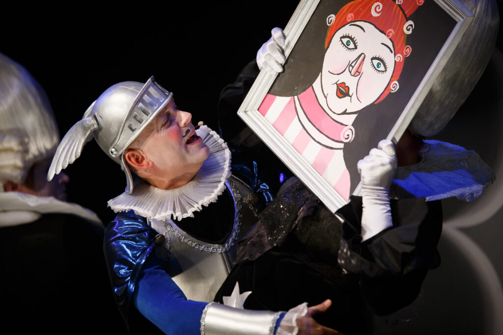 Na zdjęciu aktor w karykaturalnym przebraniu rycerza trzyma karykaturalny portret.