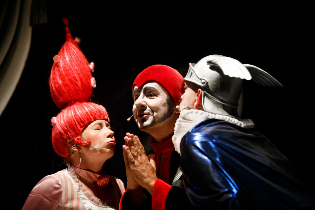 Na zdjęciu troje aktorów w abstrakcyjnych strojach w kolorach czerwonego, białego i niebieskiego. Jeden z nich ma postawione na sztorc czerwone włosy