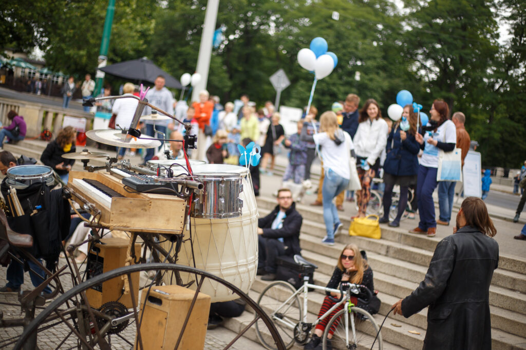 Na zewnątrz. Na zdjęciu konstrukcja przypominająca dawny rower z dużymi kołami. Do kierownicy przytwierdzone instrumenty i głośniki. W tle tłum ludzi z niebieskimi i białymi balonami Biennale