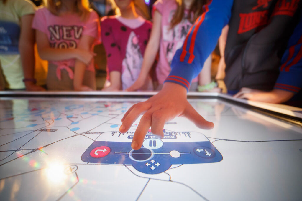 Na zdjęciu dłoń dziecka na ekranie interaktywnym. W tle inne dzieci.
