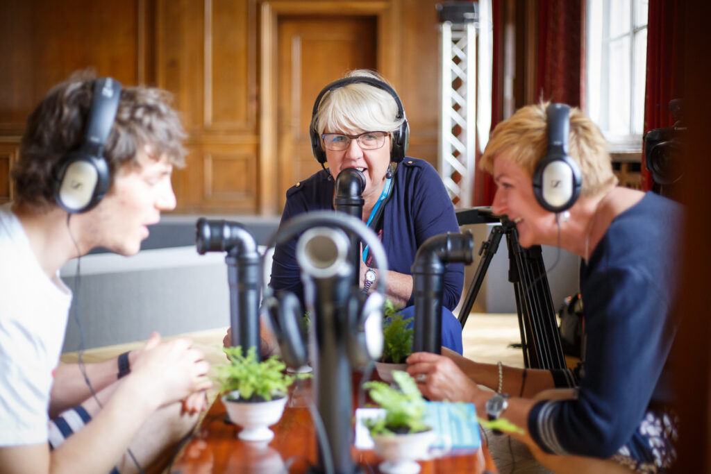 Na zdjęciu dwie starsze kobiety i mężczyzna. Wszyscy mają na uszach słuchawki i mówią coś do mikrofonów. Uśmiechają się.