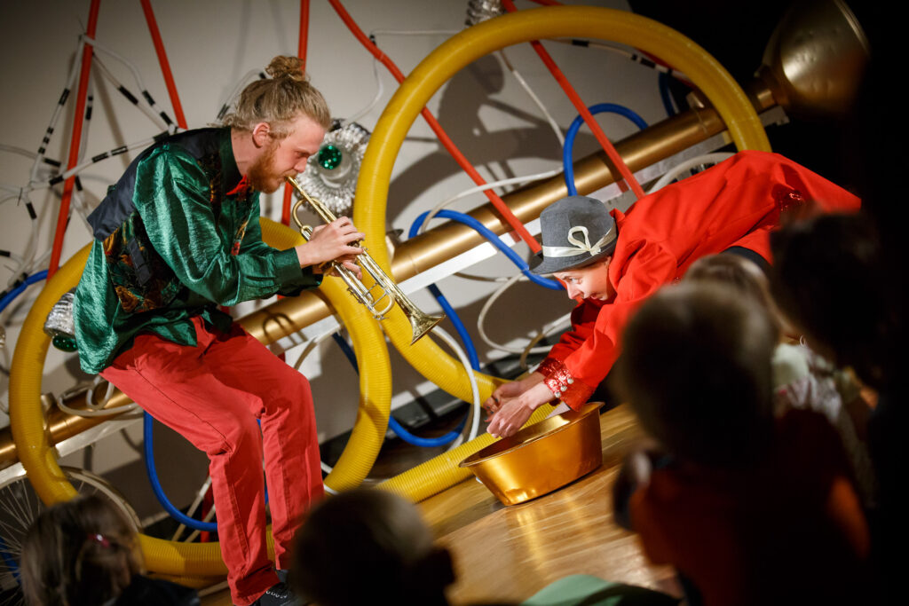 Dwoje aktorów na scenie. Aktor po lewej stronie ubrany w zieloną koszulę i czerwone spodnie gra na trąbce, a aktor po prawej stronie klęczy i trzyma dłonie wyciągnięte nad miską, która leży na podłodze. W tle kolorowa instalacja z rur.