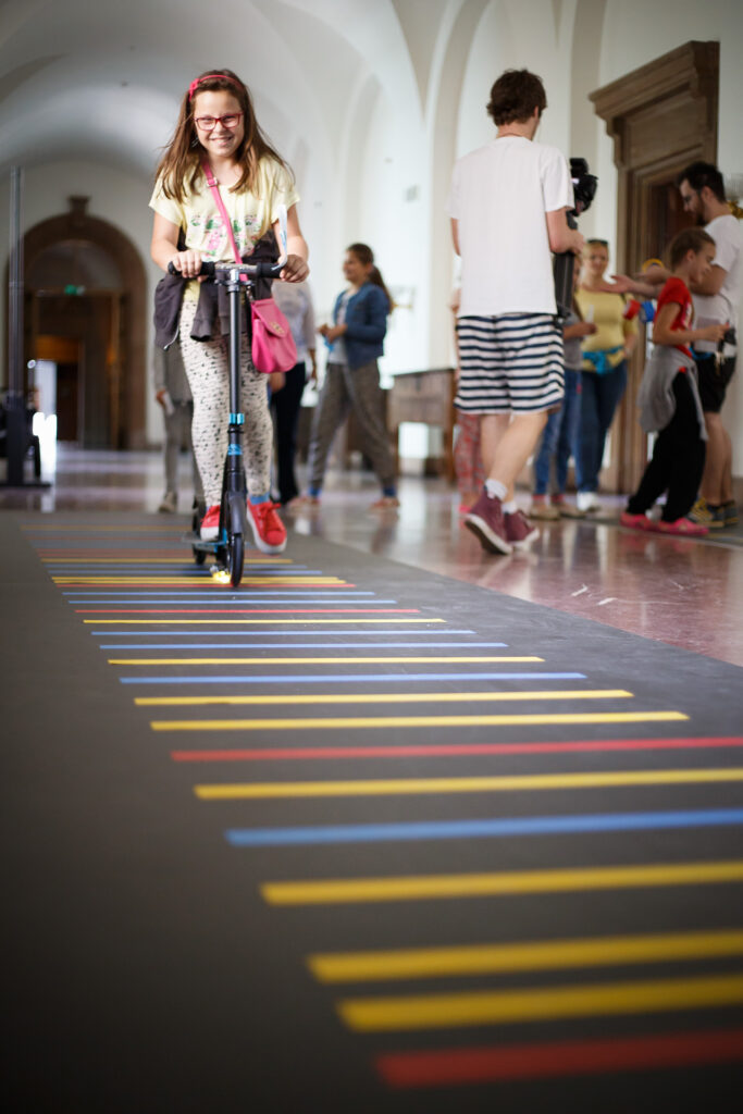 Dziewczynka jedzie na hulajnodze po kolorowej ścieżce muzycznej.